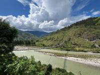 A river flows at the feet of a mountain, white clouds against blue skies hang above, a bridge is stretched across the river.